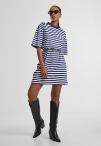 Gestreepte korte mouw jurk in marineblauw en witte katoen met een trekkoord in de taille, gecombineerd met zwarte kniehoge hakboots.