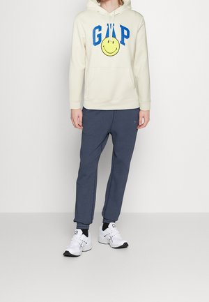 Hoodie crème avec le logo GAP bleu et un motif de smiley, associé à un pantalon de survêtement gris foncé et des baskets blanches avec des accents noirs.
