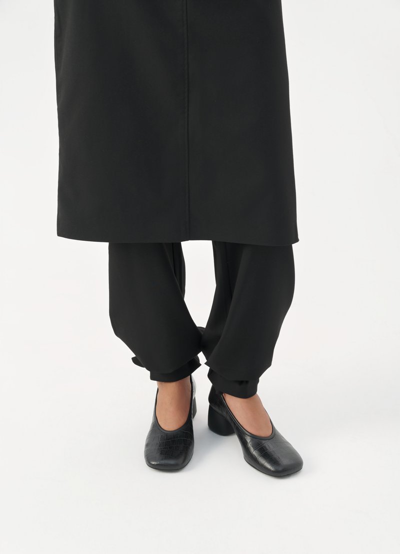 Schwarze High Heels mit strukturiertem Krokodilmuster, kombiniert mit lockeren schwarzen Hosen mit umgeschlagenen Säumen und einem schwarz geschnittenen Oberteil darüber.