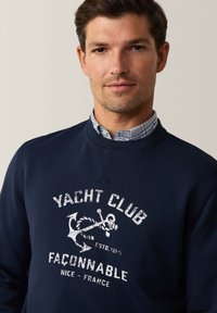 Felpa navy con scollatura a girocollo, caratterizzata da testo bianco e una grafica di un'ancora. Il materiale sembra morbido; include un colletto a quadretti blu sottostante.