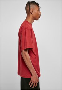 Tricou roșu din bumbac, cu o croială lejeră, mâneci scurte și decolteu rotund, prezentat din lateral pe o persoană îmbrăcată cu pantaloni scurți negri.