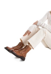 Botas marrones de cuero con cordones, con partes superiores texturizadas, cordones beige en contraste y suelas negras gruesas, llevadas con unos vaqueros blancos deshilachados.