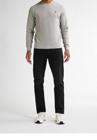 Hombre con camisa de manga larga gris claro, vaqueros negros y zapatillas blancas de pie contra un fondo blanco.