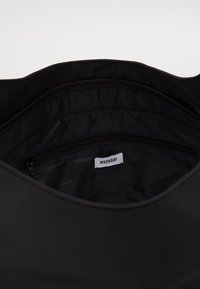 Unutrašnjost crne tkanine torbe s džepom na patentni zatvarač, glatkom teksturom i prošivenom podstavom. Unutra je vidljiv natpis "WEEKDAY".