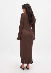 Robe en maille côtelée marron à manches longues et poignet évasé, avec une silhouette ajustée et un ourlet froncé. Portée avec des sandales à talons noires.