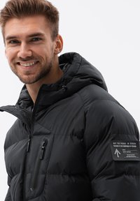 Schwarze Daunenjacke mit Kapuze, ausgestattet mit einer zippten Brusttasche, strukturiertem Stoff und einem Aufnäher am linken Ärmel mit aufgedrucktem Text.