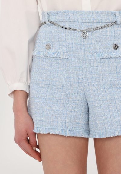 Los pantalones cortos de tweed azul claro tienen un dobladillo deshilachado, dos bolsillos y detalles decorativos en plata. Un cinturón de cadena añade un toque metálico.