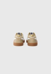 Beige og hvide sneakers med en polstret ryg, gummisål og guld-logo detalje på hælen, med glatte og strukturerede materialer.