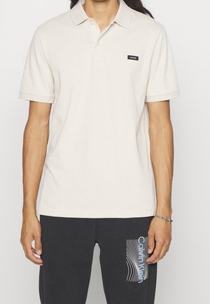 Homme portant un polo beige clair à manches courtes et un pantalon noir avec un logo Calvin Klein sur les deux vêtements.