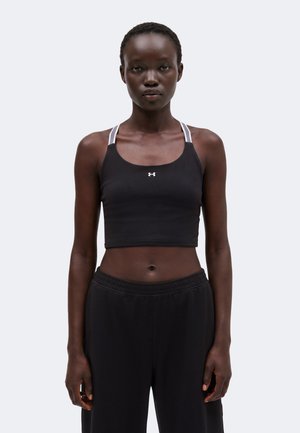 Femme portant un top de sport noir court avec des bretelles blanches et un pantalon noir ample, debout face à l'avant sur un fond uni.