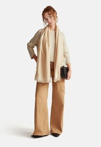 Beżowy wełniany sweter, jasnobeżowy szalik z frędzlami, brązowe szerokie spodnie i mała czarna torebka z łańcuszkiem.