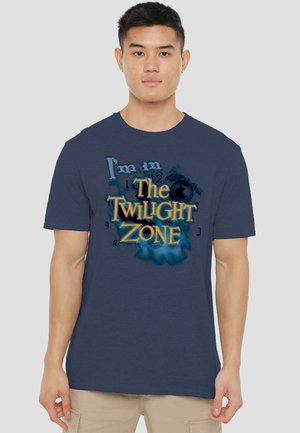 Marineblaues T-Shirt mit einem Grafikdruck, der "Ich bin in der Twilight Zone" in Gelb und Blau zeigt, gestaltet mit stilisierten Buchstaben und einem strukturierten Design.