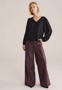 Blusa nera a maniche lunghe con scollatura a V e apertura con bottoni, abbinata a pantaloni larghi in finta pelle bordeaux con dettagli nelle cuciture.