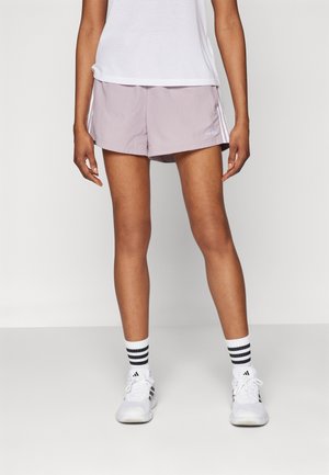 Kobieta ubrana w jasnofioletowe spodenki Adidas, biały T-shirt, białe buty Adidas oraz białe skarpetki z czarnymi paskami stojąca na jednolitym tle.