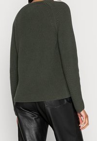 Henkilö, joka pukeutunut tummanvihreään teksturoituun neulepaitaan ja mustiin nahkahousuihin, kuvattuna takaa yksiväristä vaaleaa taustaa vasten.