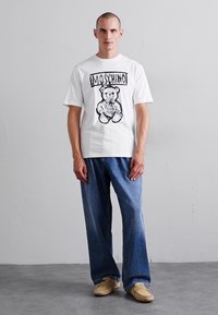 Tricou alb din bumbac cu un grafic negru al unui urs de pluș și textul "MOSCHINO." Asortat cu jeansi largi din denim albastru.