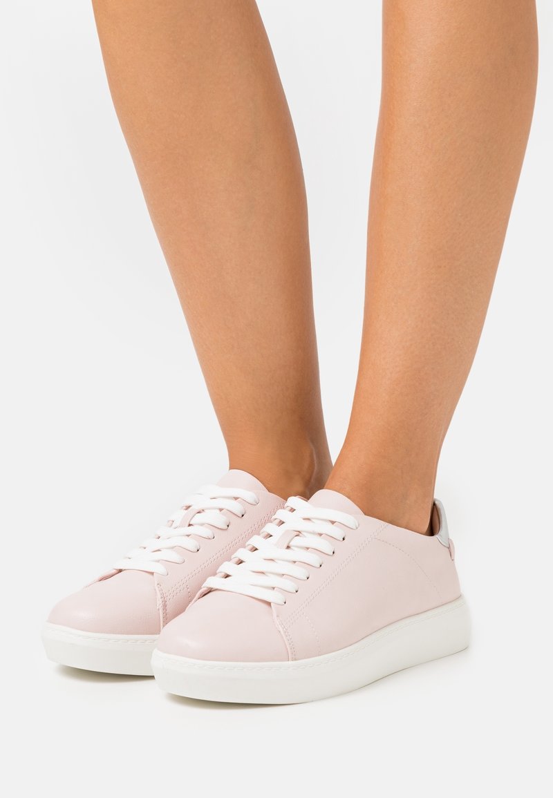 Baskets basses rose clair avec lacets blancs et semelles épaisses blanches, portées pieds nus sur un fond uni.