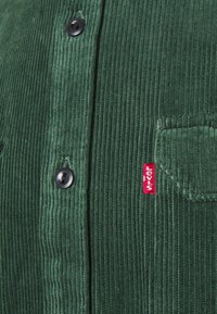 Tummanvihreä corduroypuku mustilla napilla ja pieni punainen Levi's-lappu taskussa.