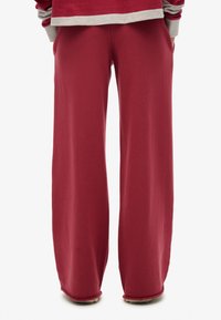 Person trägt lockere, weit geschnittene dunkelrote Hosen und ein rot-graues gestreiftes Langarmshirt, steht mit den Händen in den Taschen.