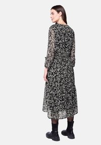 Zwart langslip-jurk met een bloemenpatroon, doorschijnende stof, smock taille en een lengte tot halverwege de kuit. Gecombineerd met zwarte enkellaarsjes.