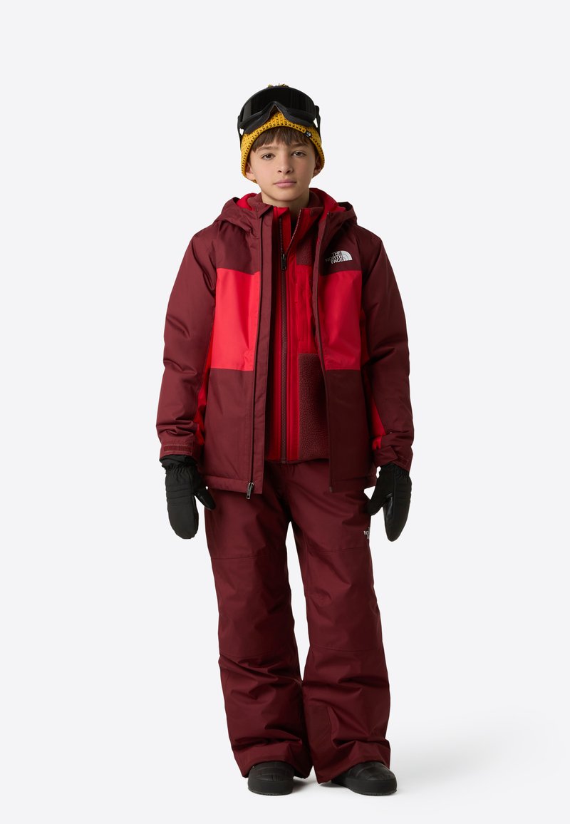 Red and maroon insulated ski jacket with a hood, paired with matching pants. Black gloves and goggles, with a yellow beanie.