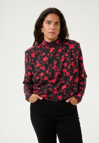 Schwarze Bluse mit langen Ärmeln und Blumenmuster in Rot. Hoher Kragen und lockerer Schnitt. Wird mit schwarzen Hosen getragen.
