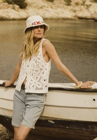 Gehaakte witte vest met bloemenpatronen, gecombineerd met lichtdenimshorts en een crèmekleurige bucket hat met rode borduursels. Natuurlijke omgeving aan het water.
