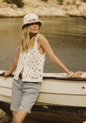 Gilet en crochet blanc avec des motifs floraux, associé à un short en denim clair et un chapeau seau crème orné de broderies rouges. Décor naturel au bord de l'eau.