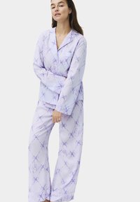 Frau, die ein helllilanes Satin-Pyjama-Set mit einer zarten Schleife und einem Wirbelmuster trägt, steht mit einem Arm verschränkt vor einem schlichten Hintergrund.