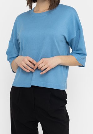 Kobieta ubrana w luźny, jasnoniebieski sweter dziany z rękawami do łokci i czarne spodnie na jednolitym tle.