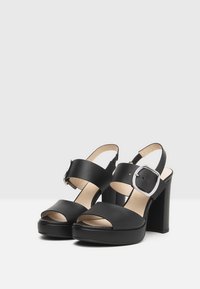 Black platform sandals with wide straps, a rounded toe, and an oversized silver buckle. Features a block heel and smooth faux leather texture.
