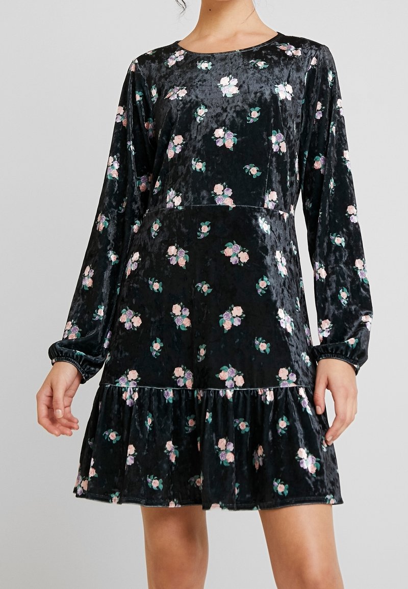Svart sammet klänning med långa puffärmar, blommönster i rosa och grönt, volangkant nertill, rund halsringning. Mjuk textur och avslappnad passform.