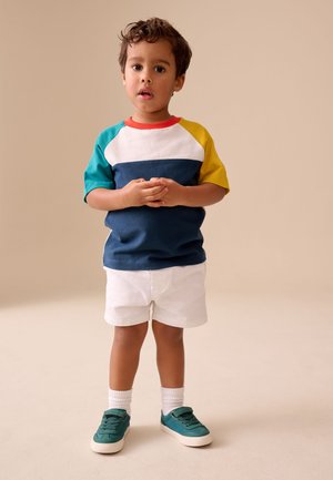 Ung dreng står med hænderne foldet, iført en flerfarvet t-shirt, hvide shorts, hvide sokker og turkis sneakers på beige baggrund.