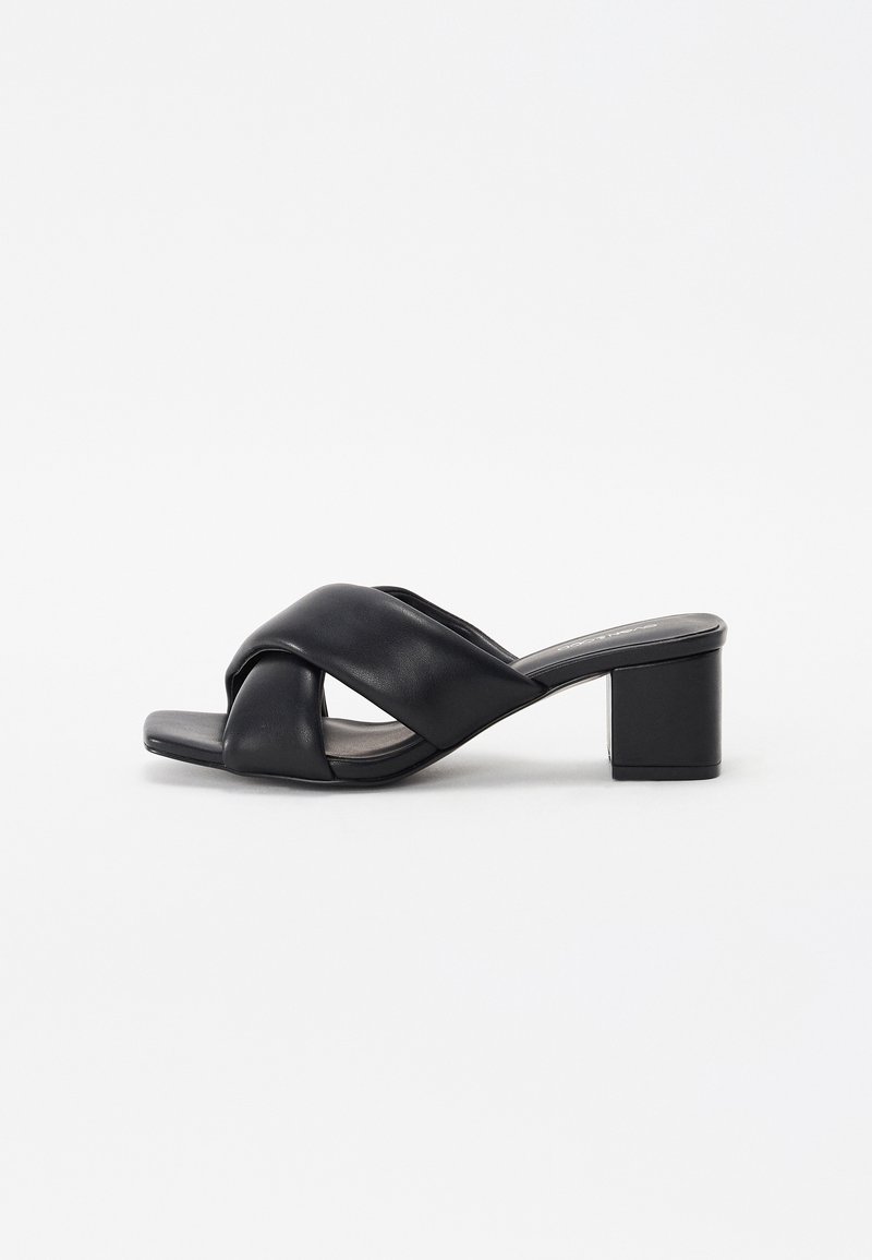 Sandale mule en cuir noir à bout ouvert, avec de larges brides croisées et un talon bloc moyen sur fond blanc.