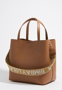 Tan leather tote bag with two top handles, featuring a detachable strap with "BIMBA Y LOLA" text. Smooth texture, minimalist design.