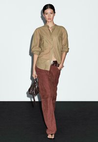 Lichtbruine, knoopsluiting shirt met lange mouwen; gecombineerd met donkerbruine suède broek; model houdt een kleine bruine tas vast; minimalistische achtergrond.