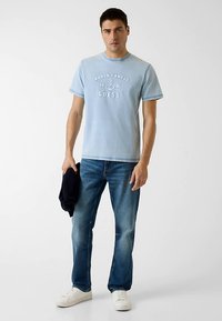 T-shirt en coton bleu clair avec l'inscription "World Famous Guess", associé à un jean en denim bleu et des baskets blanches. Le mannequin tient une veste sombre.