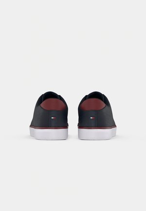 Paire de baskets basses bleu marine avec semelles blanches et accents marron au talon, vues de dos sur un fond clair uni.