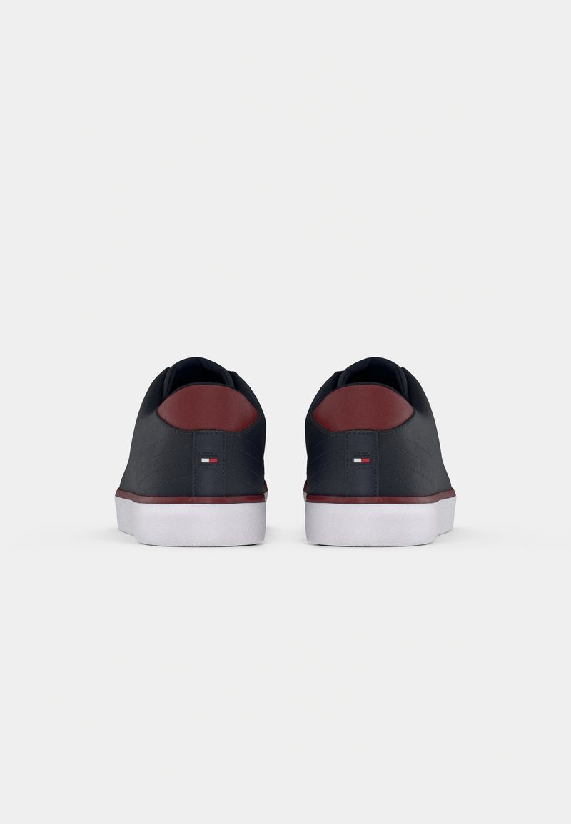 Paire de baskets basses bleu marine avec semelles blanches et accents marron au talon, vues de dos sur un fond clair uni.