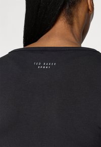 Top sport negru cu un decolteu larg și o textură fină. Logo-ul "TED BAKER SPORT" imprimat pe spate cu litere albe.