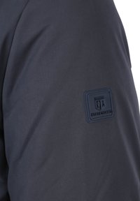 Marineblauwe jas van gladde stof, met een textuur logo patch op de bovenarm. Het ontwerp is modern en minimalistisch zonder opvallende kenmerken.