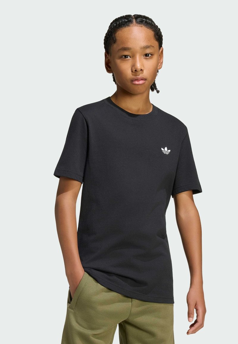 Αγόρι με πλεξούδες φοράει μαύρο μπλουζάκι Adidas και πράσινο παντελόνι με το ένα χέρι στην τσέπη, στέκεται μπροστά σε απλό φωτεινό φόντο.