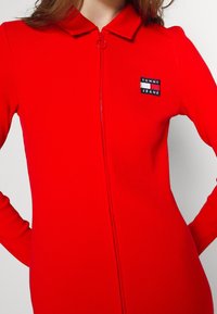 Combinaison longue à manches longues rouge côtelée avec fermeture éclair sur le devant et col pointu. Présente un patch logo Tommy Jeans sur la gauche de la poitrine.