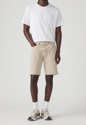 Mand iført hvid T-shirt, beige denimshorts, hvide crew-sokker og hvide og sorte løbesko, stående med den ene hånd i lommen.