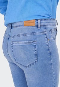 Vaqueros de mezclilla azul claro con una textura suave, que presentan bolsillos traseros y una etiqueta de cuero marrón claro que dice "ONLY CARMAKOMA" cerca de la cintura.