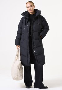 Manteau puffer noir long avec capuche, doté d'une fermeture éclair, de deux poches avant et d'une texture brillante. Accompagné d'un sac beige clair.