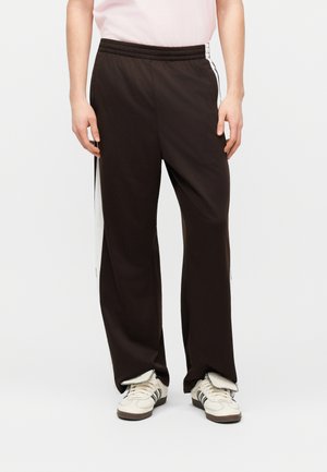 Άτομο που φοράει μαύρο φαρδύ παντελόνι φόρμας με άσπρες πλαϊνές ρίγες και λευκά αθλητικά παπούτσια με μαύρες λεπτομέρειες, στέκεται μπροστά σε απλό φόντο.