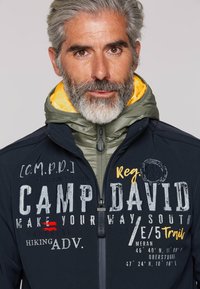 Marineblaue Jacke mit einer grünen Kapuze und geliner Fütterung. Enthält verschiedene Textgrafiken in kontrastierenden Farben. Verfügt über einen Reißverschluss und Logoprägungen.