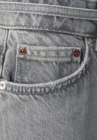 Grå denimstoff med nærbilde av en lomme, med frynsete kanter og sølvfarget maskinvare. En kobbernitt fremhever lommen.