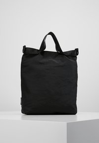 Borsa a tote nera rettangolare con manici corti e una tracolla regolabile, in posizione verticale su una superficie bianca.